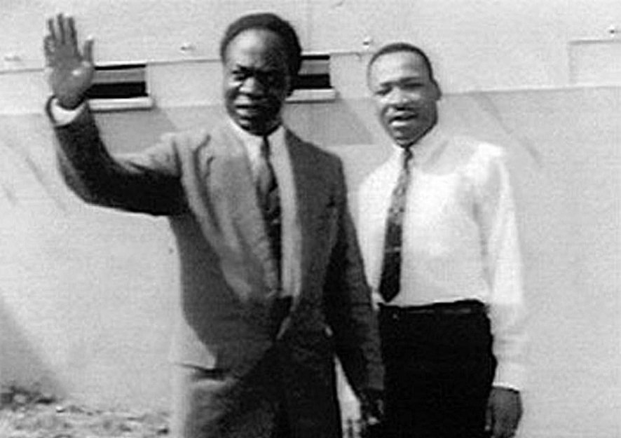 MLK and Nkrumah during a visit to Ghana, Africa in 1957. Both men stand side by side with Nkrumah in a suit and MLK in a white shirt and black tie and pants. Both are smiling while Nkrumah waves.